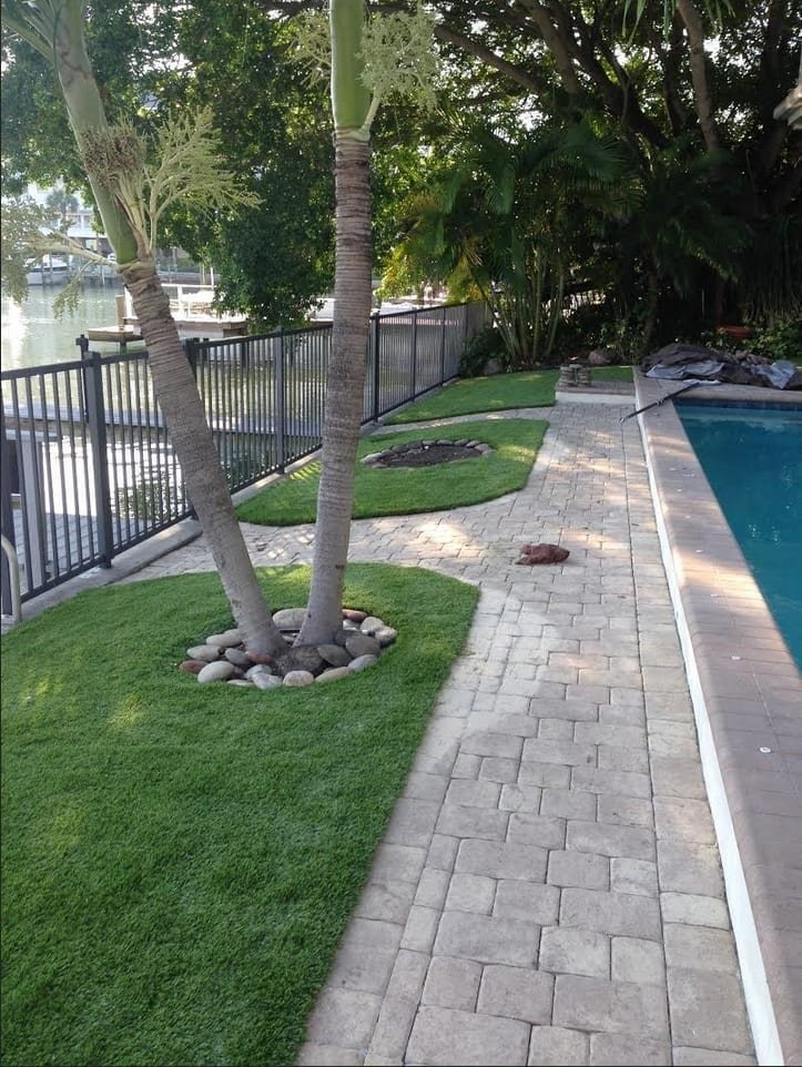 A neatly landscaped backyard with a pool, paved walkway, and grass around a tree.