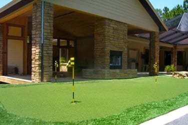 A putting green with a flagstick outside a brick house.