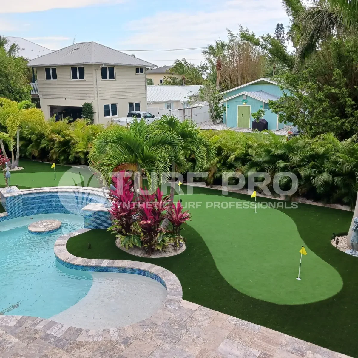 Backyard with pool and putting green surrounded by greenery and houses.